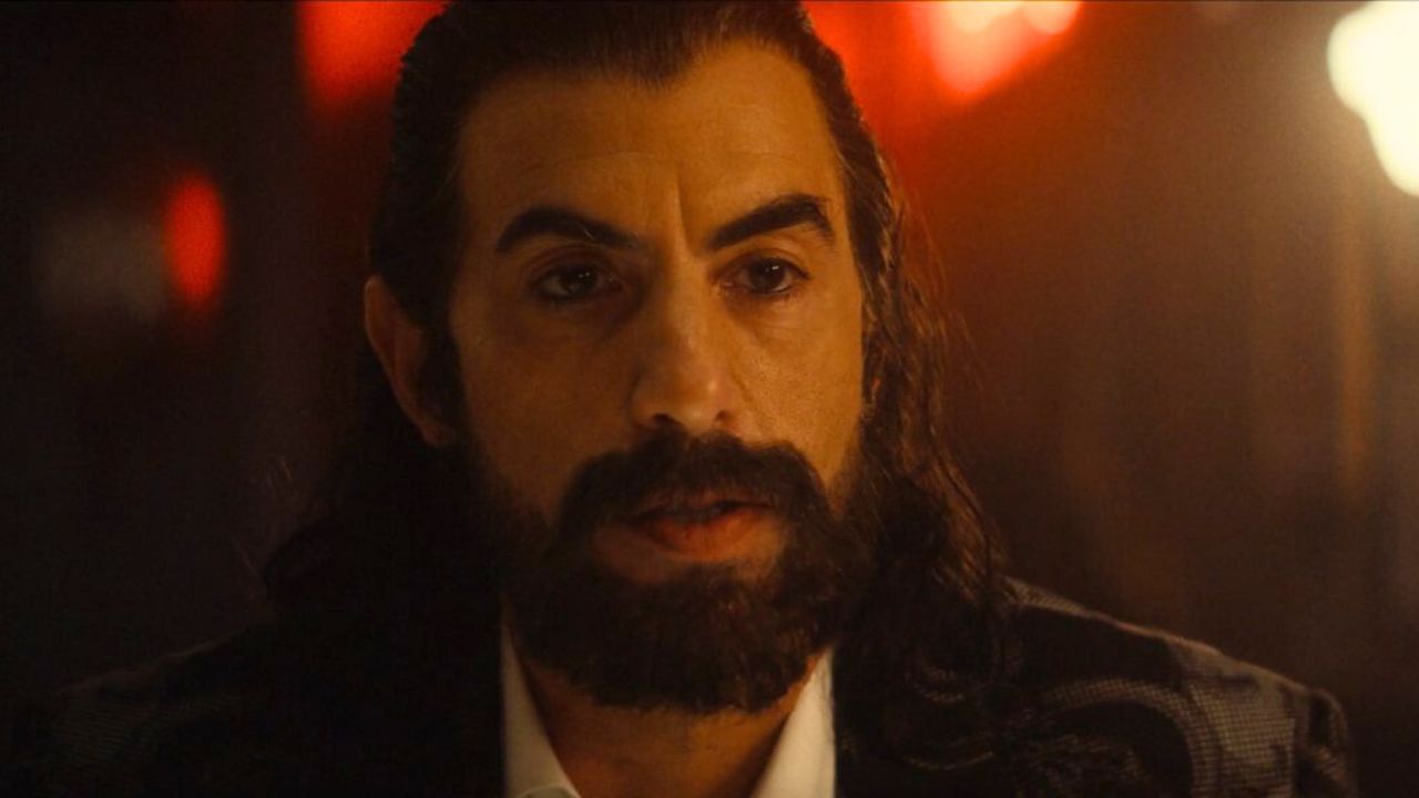 Sacha Baron Cohen as Mephisto in Ironheart, wearing a dark suit and lit by ominous red lighting in a mysterious setting.