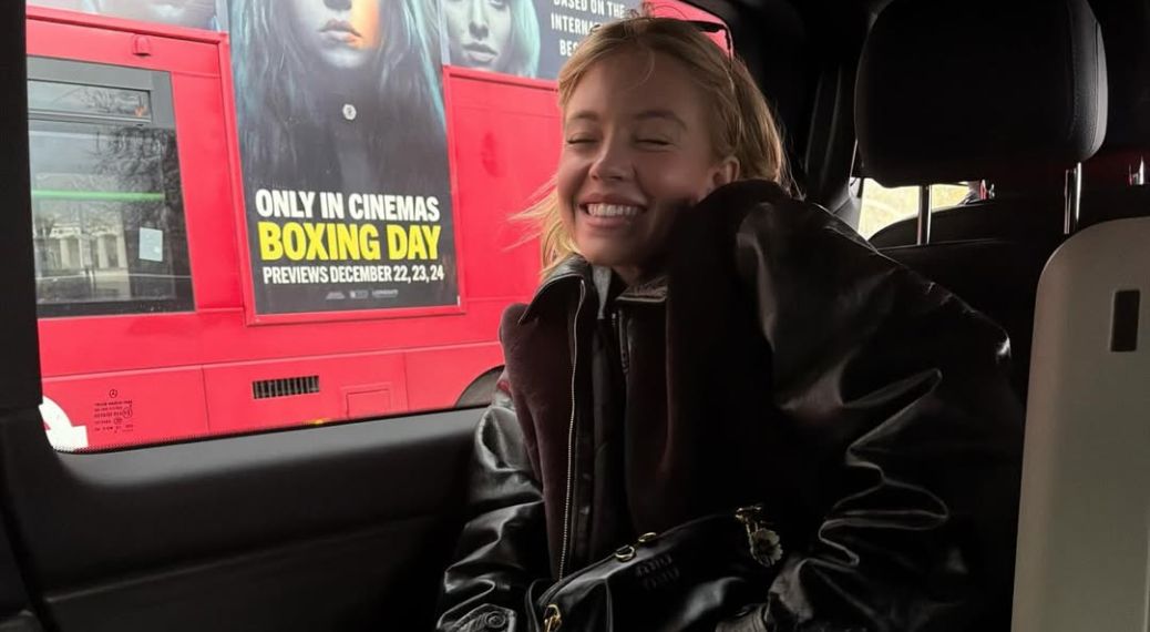 Actress Sydney Sweeney sits in the back of a vehicle, smiling with her eyes closed. She wears a dark leather jacket with a fuzzy collar and has a black Miu Miu handbag on her lap. Outside the car window, a red double-decker bus passes by, featuring a movie advertisement that reads "Only in Cinemas Boxing Day."
