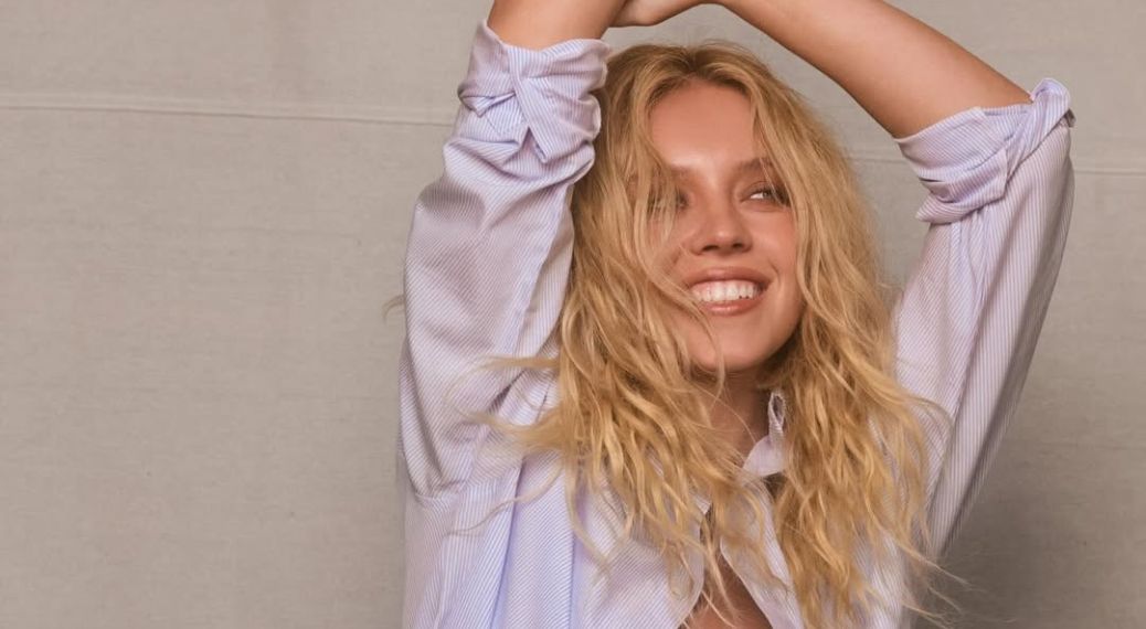 A close-up of actress Sydney Sweeney smiling broadly with her eyes slightly closed. She has long, wavy blonde hair and is wearing a light-blue and white striped button-down shirt with the sleeves rolled up. Her arms are raised above her head, and she is posed against a neutral, light-grey background.